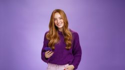 Friendly-looking relaxed and joyful nice 20s female student writing post in social network using mobile phone holding one hand in pocket and texting friend with smartphone, smiling satisfied.