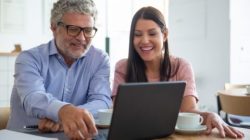 Happy cheerful mature man and young woman sitting at open laptop, looking at display, watching content over cup of coffee and laughing. Medium shot. Digital communication concept