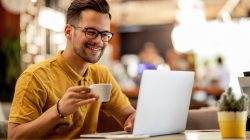 Young happy man using laptop  and drinking coffee in a cafe. Smiling man surfing the net on laptop while drinking coffee in a bar.