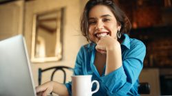Cheerful attractive young woman enjoying distant work, sitting at desk using portable computer, drinking coffee. Pretty female blogger working from home, uploading video on her channel, smiling Cheerful attractive young woman enjoying distant work, sitting at desk using portable computer, drinking coffee. Pretty female blogger working from home, uploading video on her channel, smiling