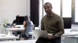 Smiling happy afro american businessman sitting on his desk and holding laptop computer in an office