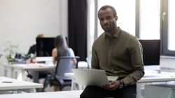 Smiling happy afro american businessman sitting on his desk and holding laptop computer in an office